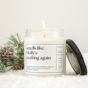May include: A white candle in a glass jar with a black lid. The candle is labeled "smells like Molly's crafting again". The label also includes the text "Infused with the scent of endless creativity, a sprinkle of colorful chaos, and the joy of handmade creations."