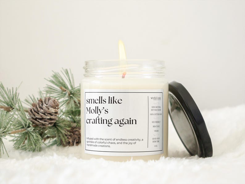 May include: A white candle in a glass jar with a black lid. The candle is labeled "smells like Molly's crafting again". The label also includes the text "Infused with the scent of endless creativity, a sprinkle of colorful chaos, and the joy of handmade creations."