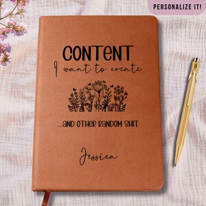 May include: A brown leather-bound journal with the text "CONTENT I want to create...AND OTHER RANDOM SHIT" in black. The journal features a floral design and the name "Jessica" in script. A gold pen rests beside the journal.