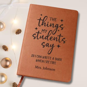 May include: A brown leather-bound journal with the text "The things my students say" in black script. The journal also includes the text "So I can write a book when I retire" and "Mrs. Johnson". A red ribbon bookmark is visible.