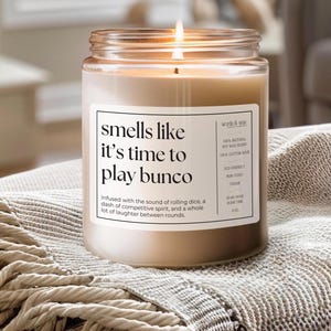 May include: A brown glass candle jar with a white label that reads "smells like it's time to play bunco". The candle is lit and the label also includes details about the candle, such as the type of wax and wick.