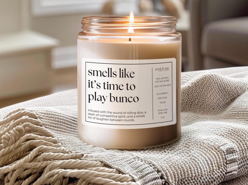May include: A brown glass candle jar with a white label that reads "smells like it's time to play bunco". The candle is lit and the label also includes details about the candle, such as the type of wax and wick.