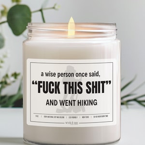 May include: A lit, glass jar candle with a white label. The label reads "a wise person once said, 'FUCK THIS SHIT' AND WENT HIKING." The candle is 9 oz and made with a natural soy wax blend. The candle has a 50-60 hour burn time.