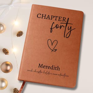 Puede incluir: Un diario de cuero marr&oacute;n con las palabras "CHAPTER forty" y un s&iacute;mbolo de coraz&oacute;n impresos en la portada. El nombre "Meredith" y la frase "each chapter holds a new adventure" tambi&eacute;n est&aacute;n impresos. Una cinta de marcador es visible.