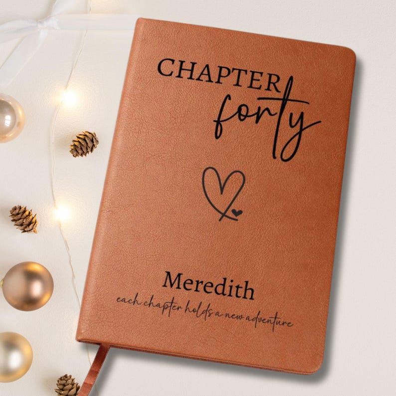Puede incluir: Un diario de cuero marr&oacute;n con las palabras "CHAPTER forty" y un s&iacute;mbolo de coraz&oacute;n impresos en la portada. El nombre "Meredith" y la frase "each chapter holds a new adventure" tambi&eacute;n est&aacute;n impresos. Una cinta de marcador es visible.