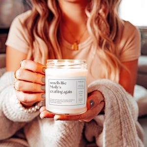 May include: A white candle with a label that reads "smells like Molly's crafting again". The candle is lit and the flame is visible. The candle is being held by a person's hand.