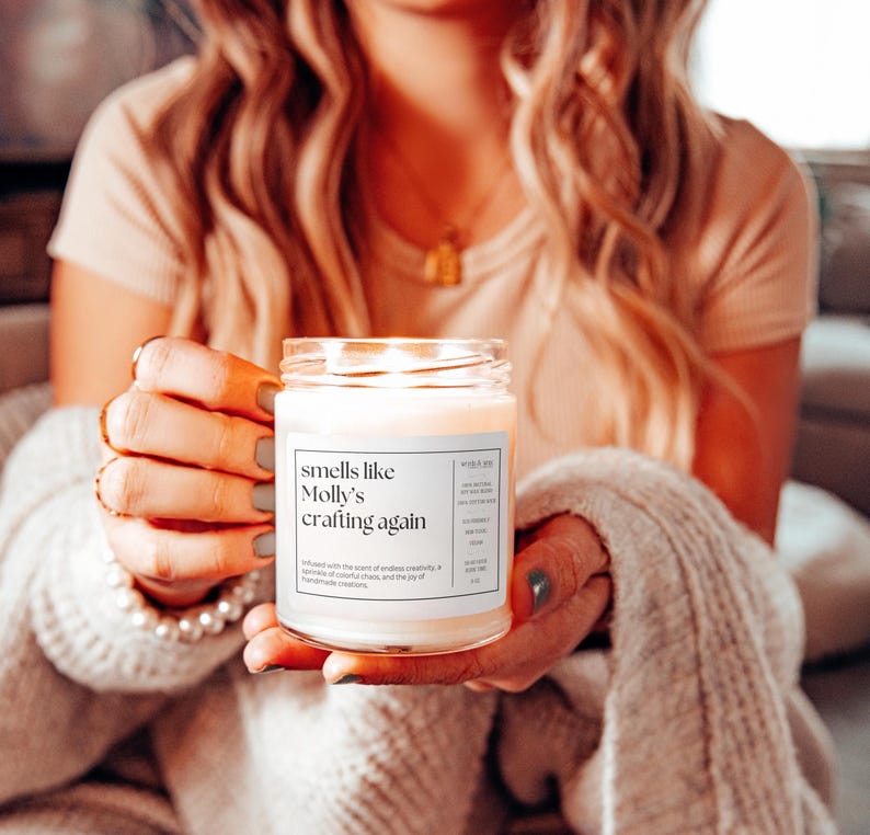 May include: A white candle with a label that reads "smells like Molly's crafting again". The candle is lit and the flame is visible. The candle is being held by a person's hand.