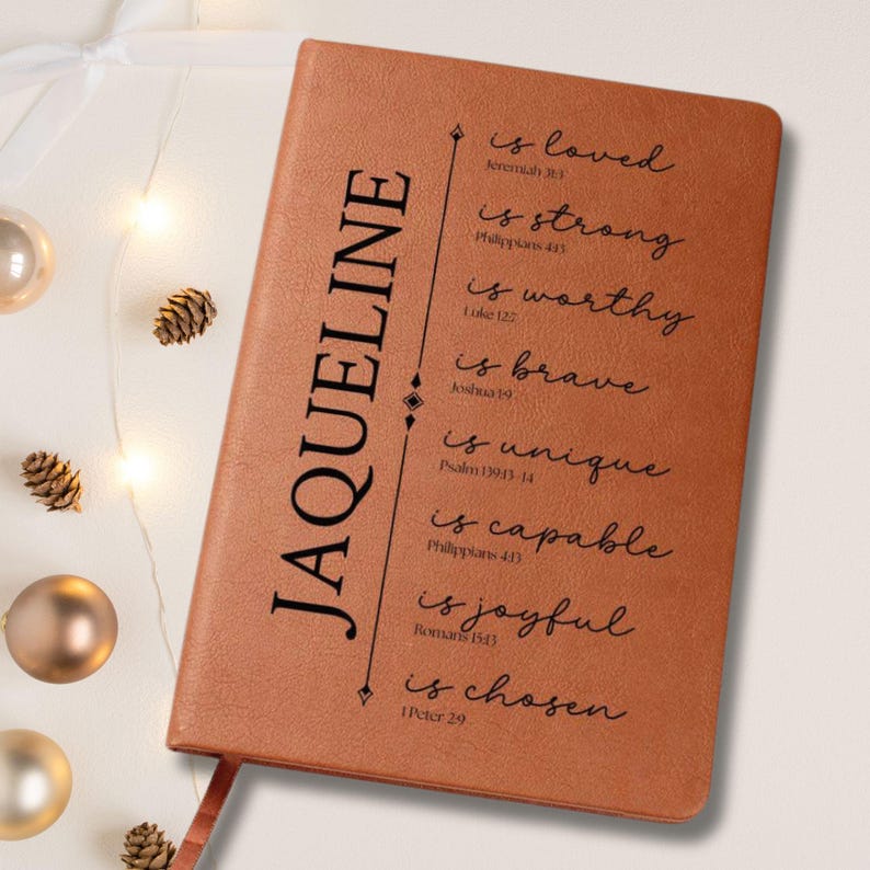 May include: A brown leather journal with the name "JAQUELINE" printed vertically on the left side. The right side features inspirational phrases like "is loved" and "is strong" in black script. A ribbon bookmark is visible.