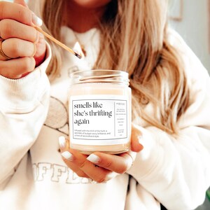 May include: A white candle with a black label that reads "smells like she's thrifting again". The candle is infused with the thrill of the hunt, a sprinkle of budget savvy brilliance, and a hint of secondhand style.