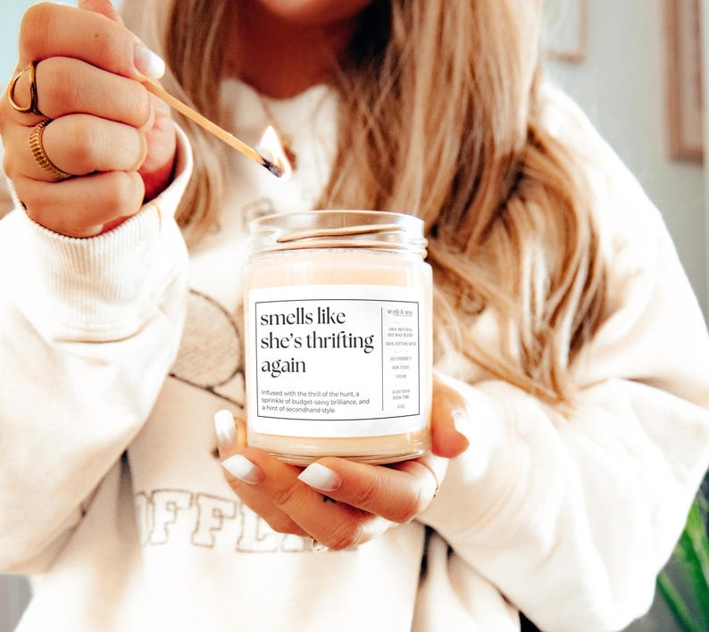 May include: A white candle with a black label that reads "smells like she's thrifting again". The candle is infused with the thrill of the hunt, a sprinkle of budget savvy brilliance, and a hint of secondhand style.