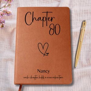 May include: A tan notebook with a leather-like texture featuring the black text Chapter 80, a heart symbol, the name Nancy, and the phrase each chapter holds a new adventure, accompanied by a gold-colored metal pen with a clip and a pointed tip.