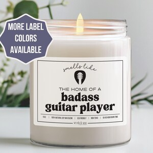May include: A white candle with a black label that reads "smells like the home of a badass guitar player".