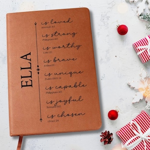 May include: A brown leather journal with the name "ELLA" printed vertically on the left side. The journal features inspirational text including "is loved," "is strong," and "is chosen." Red ornaments and wrapped gifts are in the background.