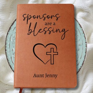 以下が含まれることがあります： 「sponsors are a blessing」の文字が黒い筆記体で書かれた茶色の革製ジャーナル。テキストの下にはハートと十字架のデザインがあり、下部には「Aunt Jenny」と印刷されています。ジャーナルは装飾的なお皿の上にあります。