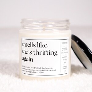 May include: A white candle in a glass jar with a black lid. The label reads "smells like she's thrifting again" and includes the text "infused with the thrill of the hunt, a sprinkle of budget-savvy brilliance, and a hint of secondhand style."
