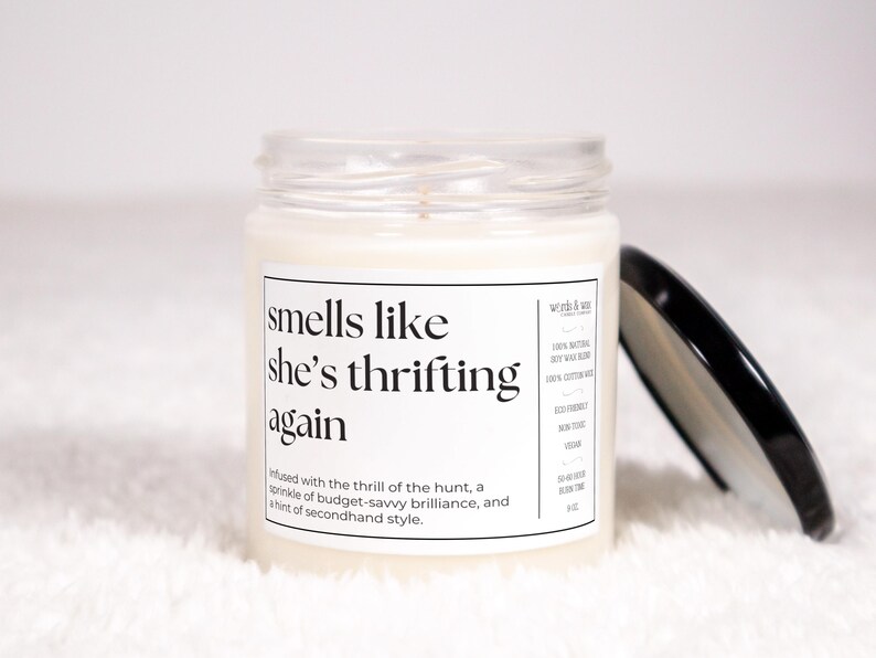 May include: A white candle in a glass jar with a black lid. The label reads "smells like she's thrifting again" and includes the text "infused with the thrill of the hunt, a sprinkle of budget-savvy brilliance, and a hint of secondhand style."