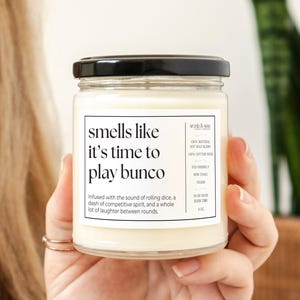 May include: A white candle in a glass jar with a black lid. The label reads "smells like it's time to play bunco" and includes the text "Infused with the sound of rolling dice, a dash of competitive spirit, and a whole lot of laughter between rounds."