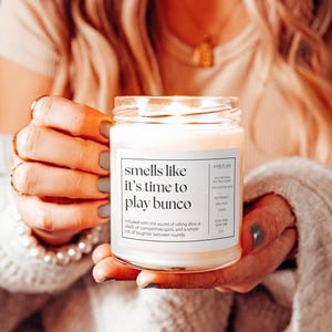 May include: A white candle in a glass jar with the text "smells like it's time to play bunco". The candle is lit and the text is surrounded by a white background.