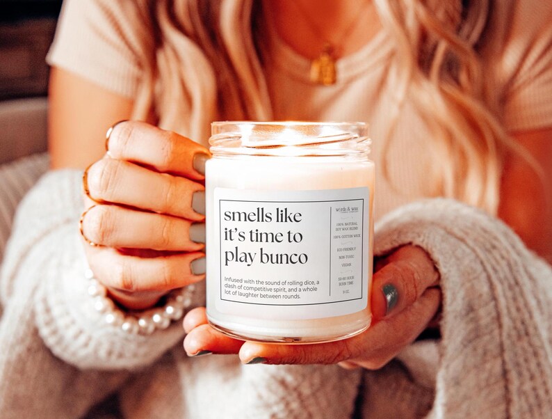 May include: A white candle in a glass jar with the text "smells like it's time to play bunco". The candle is lit and the text is surrounded by a white background.