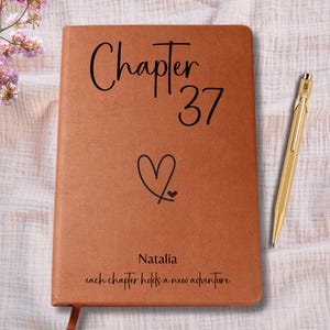 May include: A brown leather journal with the words "Chapter 37" and a heart symbol. The name "Natalia" and the phrase "each chapter holds a new adventure" are also inscribed. A gold pen rests beside the journal.