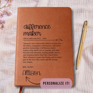 May include: A brown leather journal with the words "difference maker" and its definition printed on the cover. A gold pen rests beside the journal. The journal is personalized with the name "Allison" and the words "Personalize It!"