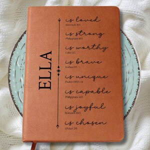 May include: A brown leather journal with the name "ELLA" printed vertically on the left side. The journal features inspirational text including "is loved," "is strong," and "is chosen." The journal is resting on a decorative plate.