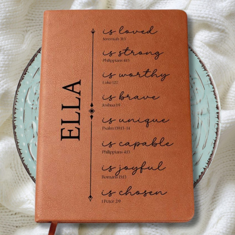 May include: A brown leather journal with the name "ELLA" printed vertically on the left side. The journal features inspirational text including "is loved," "is strong," and "is chosen." The journal is resting on a decorative plate.