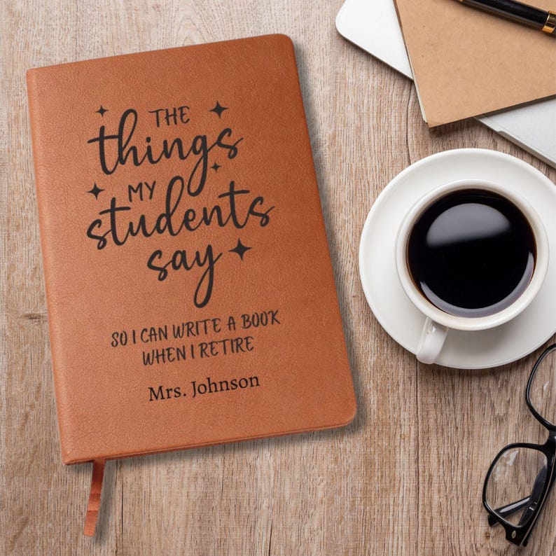 May include: A brown journal with the text "The things my students say" and "So I can write a book when I retire, Mrs. Johnson." A cup of coffee, glasses, and a laptop are on a wooden surface.