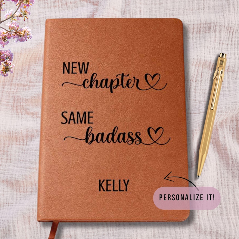 May include: A brown journal with the text "NEW chapter, SAME badass" and the name "KELLY" printed on the cover. A gold pen rests on the right side. The journal has a ribbon bookmark. A pink sticker says "PERSONALIZE IT!"