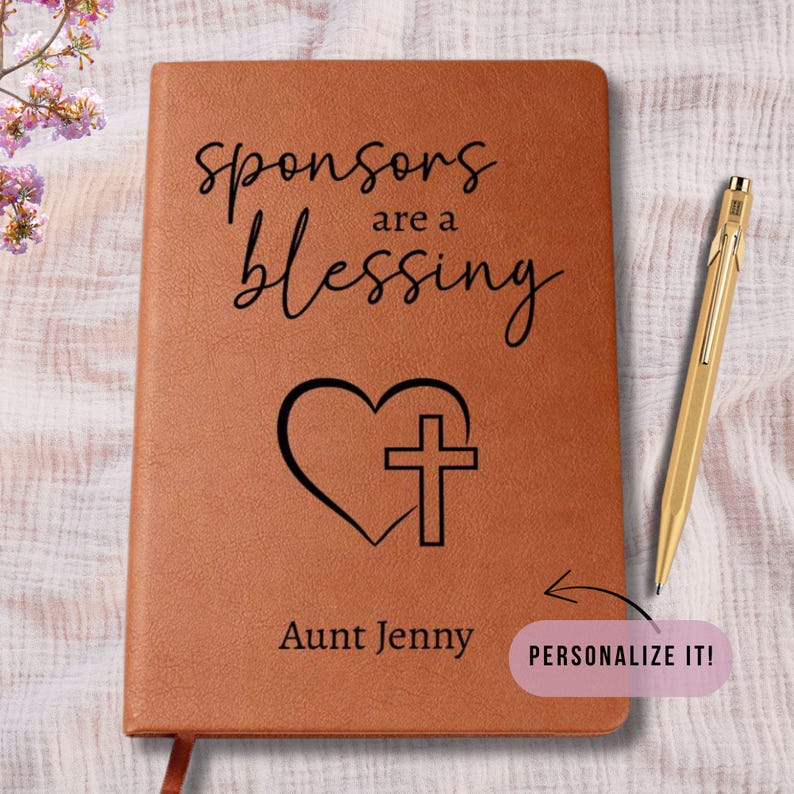 以下が含まれることがあります： 「Sponsors are a Blessing」の文字が書かれた茶色の革製ジャーナル。テキストの下にはハートと十字架のデザインが刻まれています。「Aunt Jenny」という名前が下部に印刷されています。右側には金のペンが置かれています。