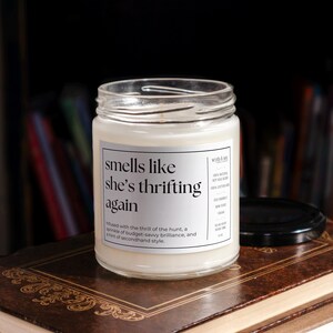 May include: A white candle in a glass jar with a label that reads "smells like she's thrifting again". The label also includes the text "Infused with the thrill of the hunt, a sprinkle of budget-savvy brilliance, and a hint of secondhand style."