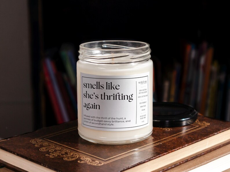 May include: A white candle in a glass jar with a label that reads "smells like she's thrifting again". The label also includes the text "Infused with the thrill of the hunt, a sprinkle of budget-savvy brilliance, and a hint of secondhand style."