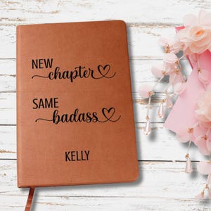 May include: A brown leather-bound journal with the text "NEW chapter" and "SAME badass" in black script. The name "KELLY" is printed at the bottom. A pink ribbon bookmark is visible. The journal is a personalized gift.