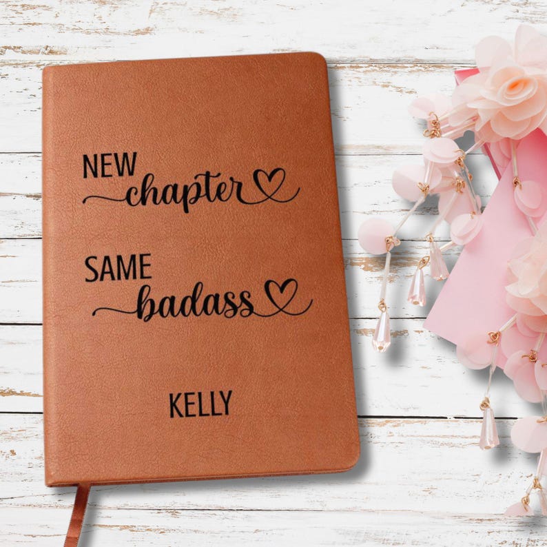 May include: A brown leather-bound journal with the text "NEW chapter" and "SAME badass" in black script. The name "KELLY" is printed at the bottom. A pink ribbon bookmark is visible. The journal is a personalized gift.