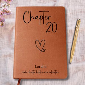May include: A brown leather journal with the words "Chapter 20" and a heart symbol. The name "Loralie" and the phrase "each chapter holds a new adventure" are also printed on the journal. A gold pen rests beside the journal.