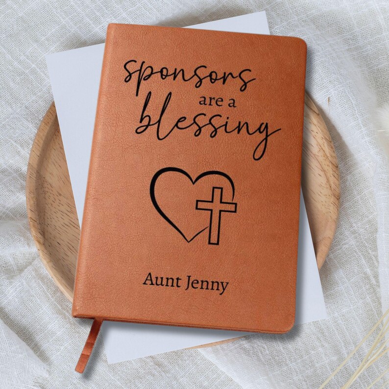 以下が含まれることがあります： 「Sponsors are a blessing」の文字が書かれた茶色の革製ジャーナル。テキストの下には、ハートと十字架が刻まれています。「Aunt Jenny」という名前が下部に印刷されています。赤いリボンブックマークが見えます。