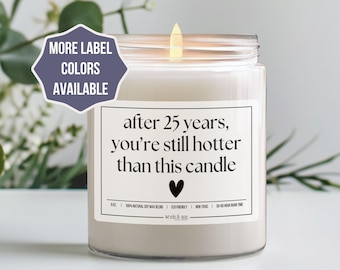 May include: A lit, white candle in a clear glass jar. The label reads "after 25 years, you're still hotter than this candle" with a black heart. A purple hexagon with text "MORE LABEL COLORS AVAILABLE" is in the upper left.
