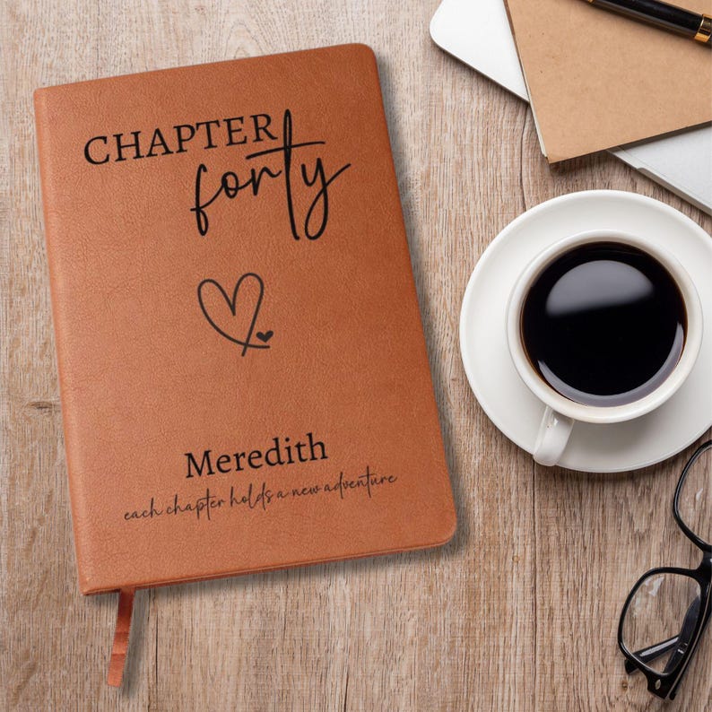 Puede incluir: Un diario de cuero marr&oacute;n con las palabras "CHAPTER forty" y un s&iacute;mbolo de coraz&oacute;n impresos en la portada. El nombre "Meredith" y la frase "each chapter holds a new adventure" tambi&eacute;n est&aacute;n impresos. Una taza de caf&eacute; y gafas est&aacute;n sobre la superficie de madera.