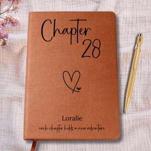 May include: A brown leather journal with the words "Chapter 28" and a heart symbol. The name "Loralie" and the phrase "each chapter holds a new adventure" are also printed on the cover. A gold pen rests beside the journal.