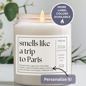 May include: A lit, white candle in a glass jar with a label that reads "smells like a trip to Paris." The label also includes the text "words & wax" and details about the candle's composition and burn time. A round sticker says "More label colors available."