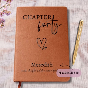 May include: A brown leather journal with the words "CHAPTER forty" and a heart symbol. The name "Meredith" is below the heart, with the phrase "each chapter holds a new adventure". A gold pen rests beside the journal.
