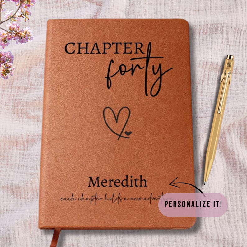 Puede incluir: Un diario de cuero marr&oacute;n con las palabras "CHAPTER forty" y un s&iacute;mbolo de coraz&oacute;n. El nombre "Meredith" est&aacute; debajo del coraz&oacute;n, con la frase "cada cap&iacute;tulo contiene una nueva aventura". Un bol&iacute;grafo dorado descansa junto al diario.