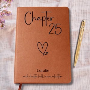May include: A brown leather journal with the text "Chapter 25" and a heart symbol. The name "Loralie" and the phrase "each chapter holds a new adventure" are also printed on the cover. A gold pen rests beside the journal.