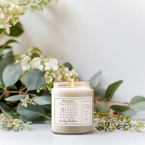 May include: A white candle with a clear glass jar and a label that says "March 2023" and has a calendar printed on it. The label also says "one day at a time".