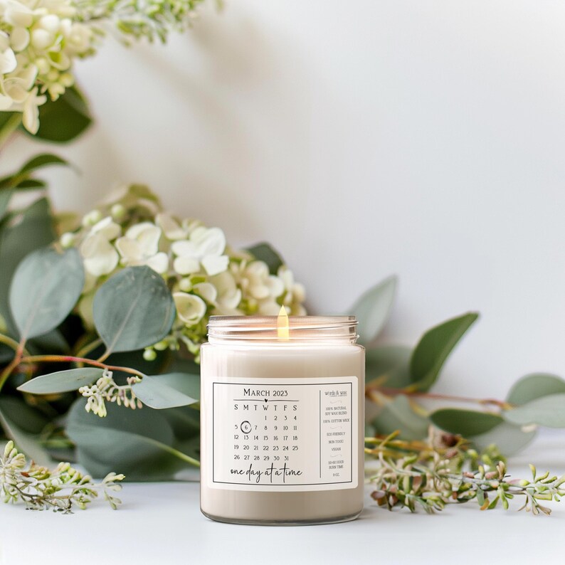 May include: A white candle with a clear glass jar and a label that says "March 2023" and has a calendar printed on it. The label also says "one day at a time".
