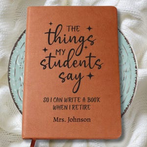 May include: A brown leather-bound journal with the text "The things my students say" in black script. The journal also includes the text "So I can write a book when I retire" and "Mrs. Johnson". The journal has a ribbon bookmark.