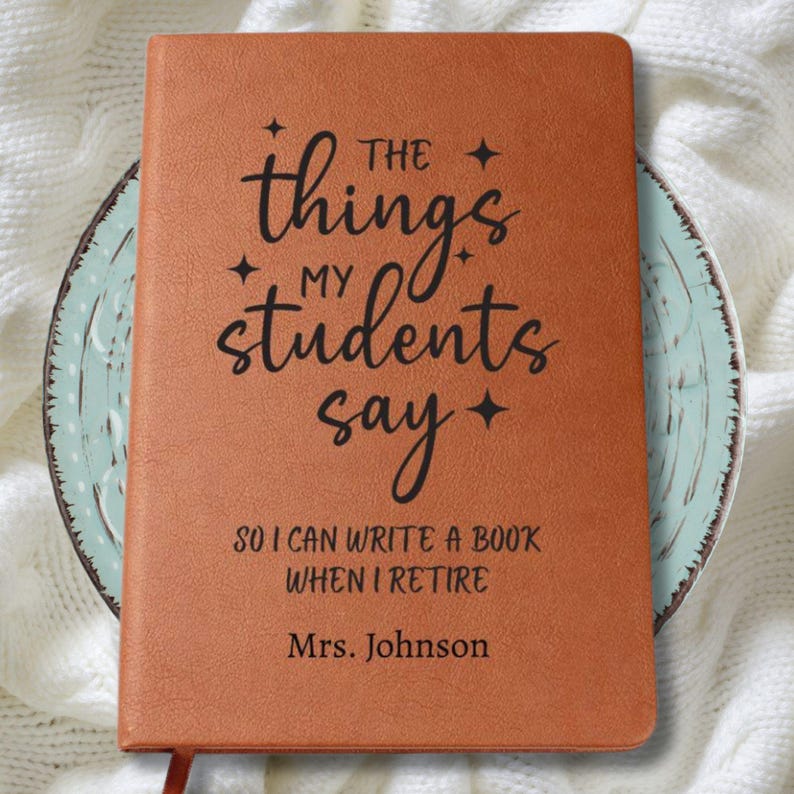 May include: A brown leather-bound journal with the text "The things my students say" in black script. The journal also includes the text "So I can write a book when I retire" and "Mrs. Johnson". The journal has a ribbon bookmark.