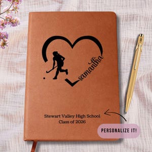 May include: A brown leather-bound notebook with a black heart design featuring a field hockey player silhouette and the name "Samantha." The notebook also includes the text "Stewart Valley High School Class of 2026" and a gold pen. The text "PERSONALIZE IT!" is also visible.