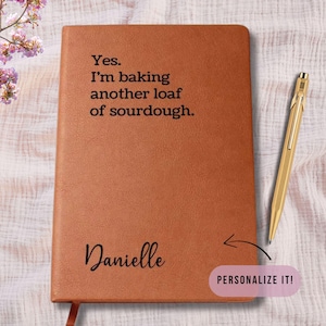 May include: A brown leather journal with the text "Yes. I'm baking another loaf of sourdough." and the name "Danielle" in script. A gold pen rests on the right side. A pink "Personalize It!" sticker is in the lower right corner.