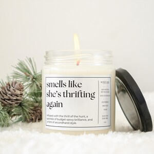May include: A white candle in a glass jar with a black label that reads "smells like she's thrifting again". The label also includes the text "Infused with the thrill of the hunt, a sprinkle of budget-savvy brilliance, and a hint of secondhand style."